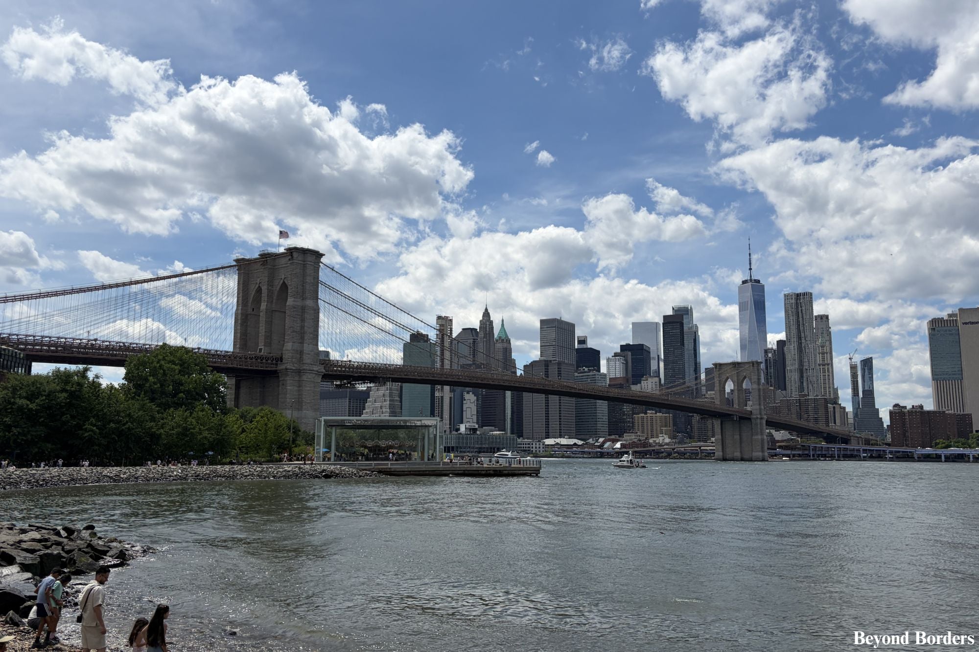The Lower Manhattan skyline featuring the Brooklyn Bridge and One World Trade Center under a cloudy blue sky, captured from DUMBO in Brooklyn, across the East River.