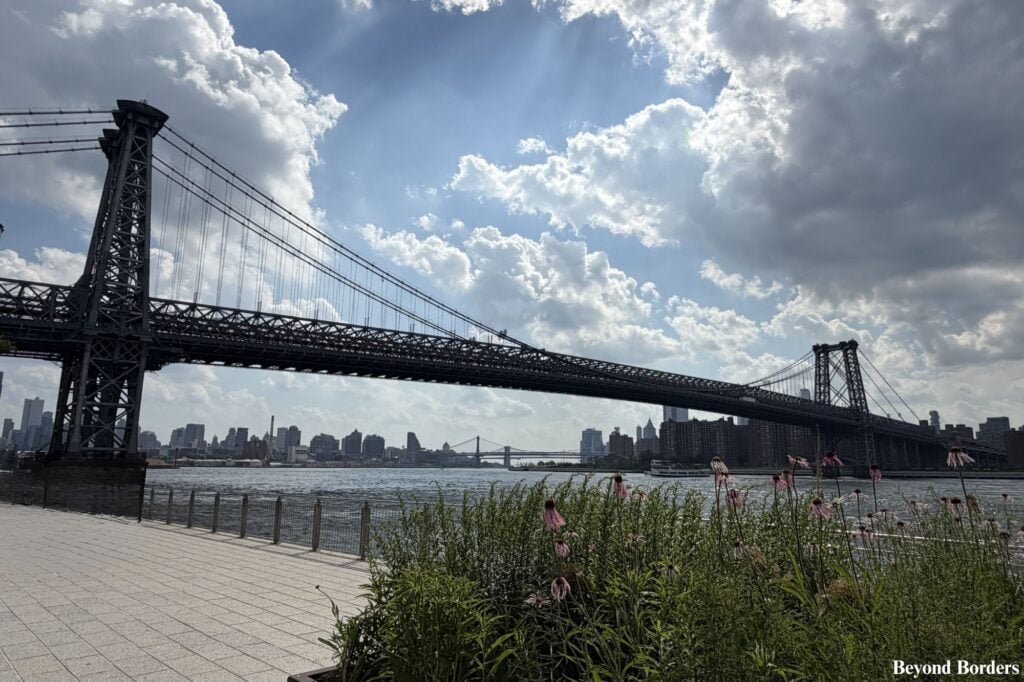 Wide view of the industrial pink steel of the Williamsburg Bridge crossing the East River from the waterfront at Domino Park in Brooklyn
