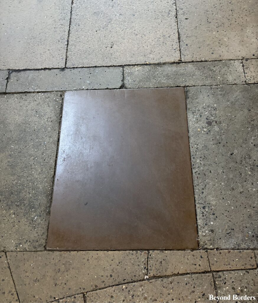The opaque panel a few steps to the left of the brass map in front of 85 Broad Street in Manhattan, NYC; people used to look through the window down at the actual excavated stone ruins of the 1670 Lovelace Tavern buried beneath the modern city